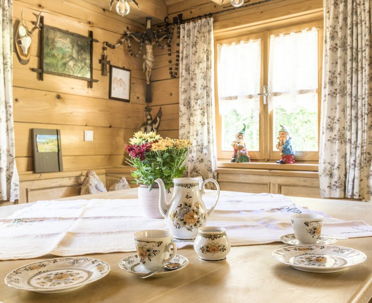 Einladend gedeckter Holztisch in der gemütlichen Stube der Almhütte, verziert mit traditionellem Kaffeegeschirr und bunten Blumen.