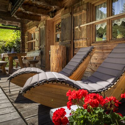 Die einladende Holzterrasse der Almhütte mit gemütlichen Sonnenliegen und bunten Geranien.