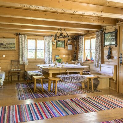 Die gemütliche Stube der Almhütte mit einem einladenden Holztisch, rustikalen Bänken mit Fellauflagen und bunten Webteppichen.