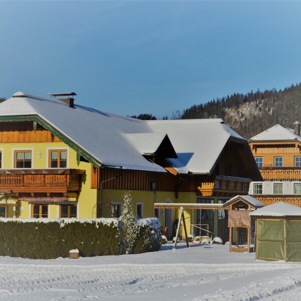 Der Bauernhof im Winter mit schneebedeckten Dächern, Holzbalkonen und einem Spielplatz für Kinder.