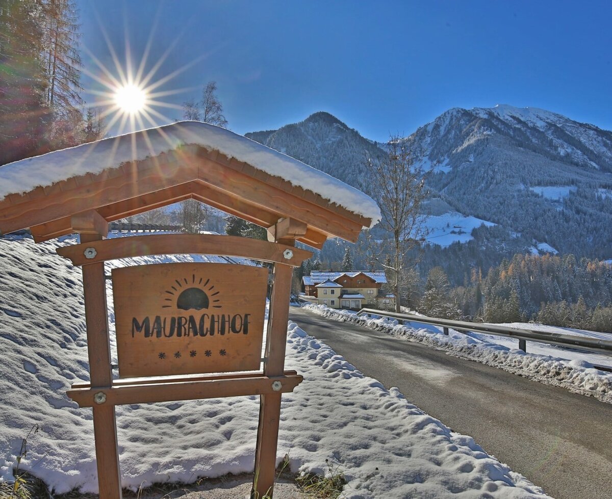 Das einladende, hölzerne Schild des Bauernhofs Maurachhof in einer sonnigen, verschneiten Winterlandschaft mit Blick auf die Alpen.