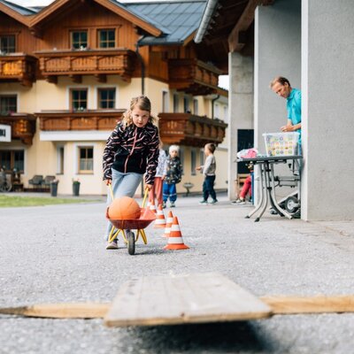 Ein lebhafter Spielbereich auf dem Bauernhof, wo Kinder mit Spielzeug-Schubkarren und Hütchen einen kleinen Parcours bewältigen können.