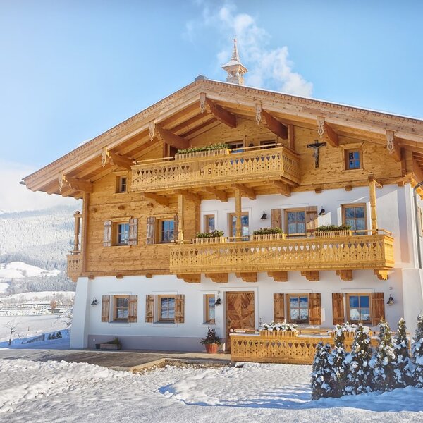 Die Fassade des Bauernhofs mit seinen charakteristischen Holzbalkonen und Holzläden, eingebettet in eine verschneite Winterlandschaft.