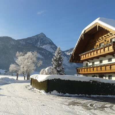 Der Bauernhof im Winter mit traditionellen Holzbalkonen, schneebedecktem Dach und einer verschneiten Berglandschaft im Hintergrund.