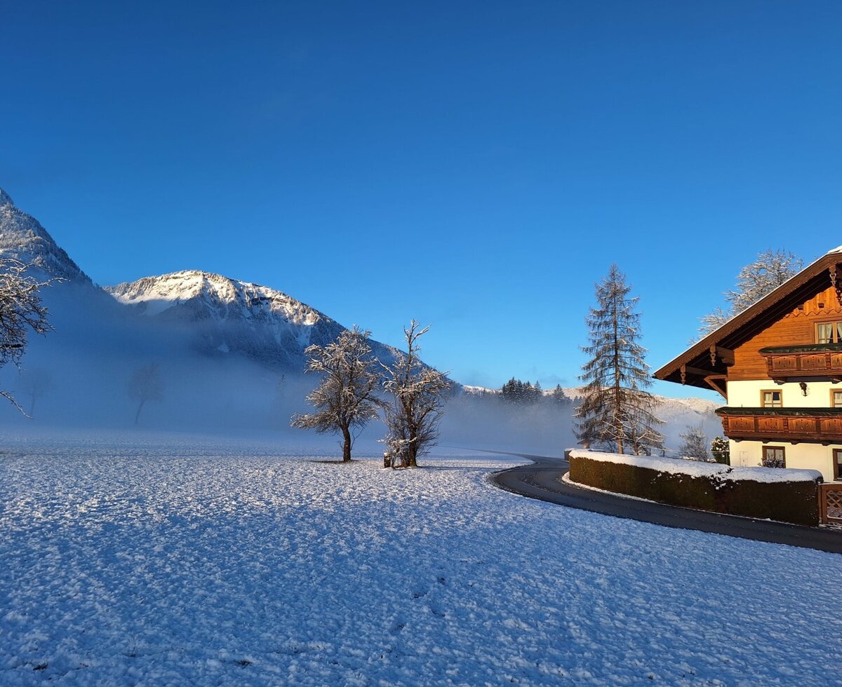 Der Bauernhof im Winter mit schneebedeckten Feldern und Bergen im Hintergrund unter blauem Himmel.
