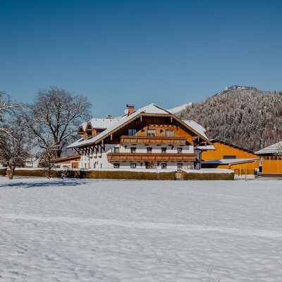 Der Bauernhof im Winter, mit schneebedeckten Dächern und Feldern, umgeben von Bergen.