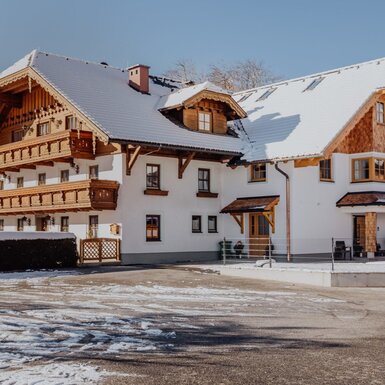 Der Bauernhof im Winter mit verschneitem Dach und Holzbalkonen vor einer Bergkulisse.