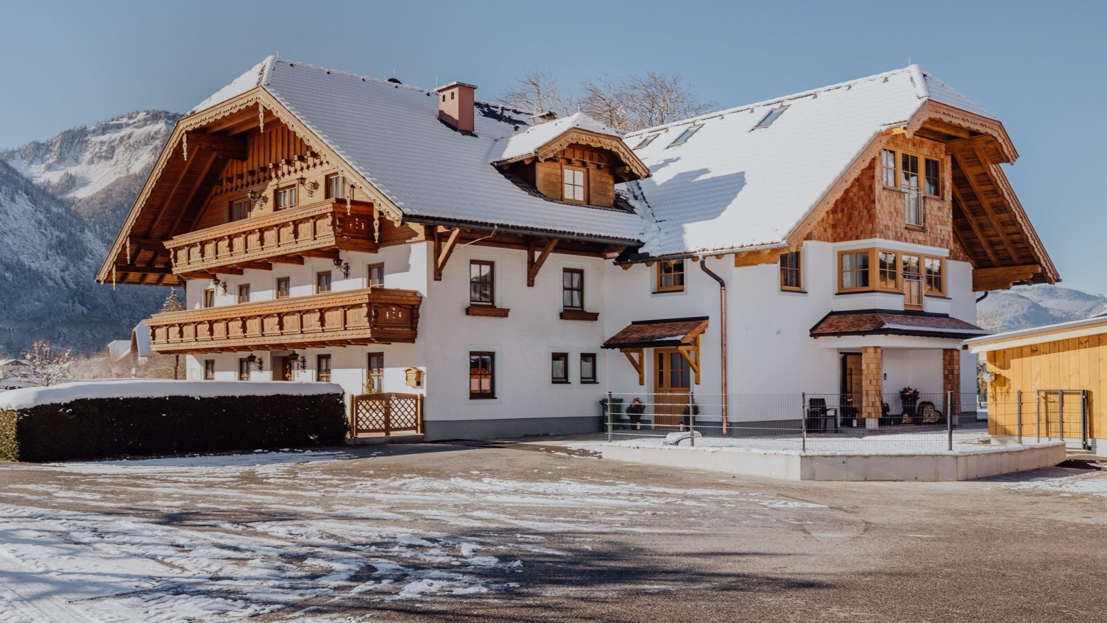 Der Bauernhof im Winter mit verschneitem Dach und Holzbalkonen vor einer Bergkulisse.