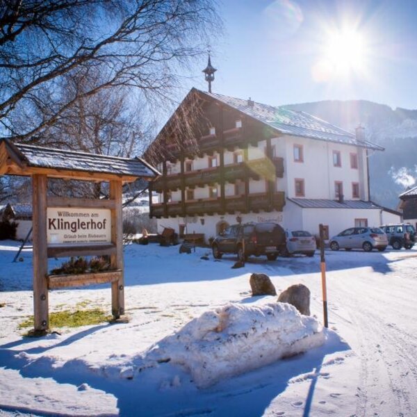 Das Bauernhofgebäude im Winter, mit dem Willkommensschild, Parkplätzen und den umliegenden Bergen.