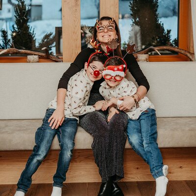 Eine Familie mit Mutter und zwei Kindern sitzt auf einer Bank im Bauernhof, alle tragen festliche Accessoires.