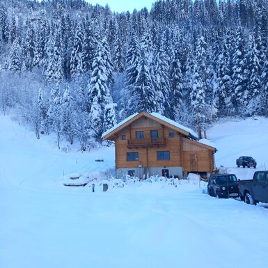 Der Hof als Holzhaus in einer tief verschneiten Winterlandschaft, umgeben von Tannen und mit Parkmöglichkeiten.