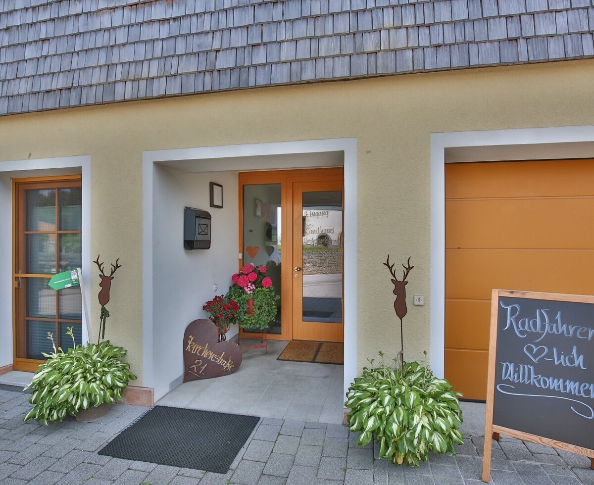 Eingang mit Holztür, Pflanzen und einem Schild, das Radfahrer willkommen heißt.