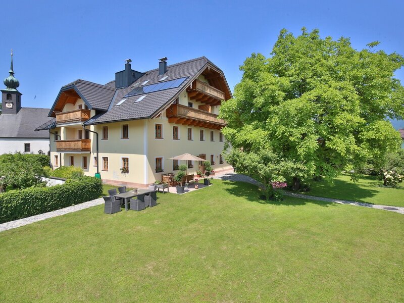 Das Bauernhofgebäude mit Balkonen und Solarpaneelen, einem Garten mit Rasenfläche und Sitzgelegenheiten im Freien, sowie einer Kirche im Hintergrund.