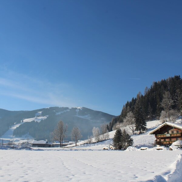 Der Bauernhof im Winter, mit einer verschneiten Landschaft und Bergen mit Skipisten im Hintergrund.