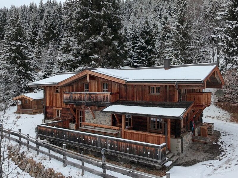 Die verschneite Alm von außen, ein Holzgebäude mit Balkonen, umgeben von Tannen und einem Holzzaun.
