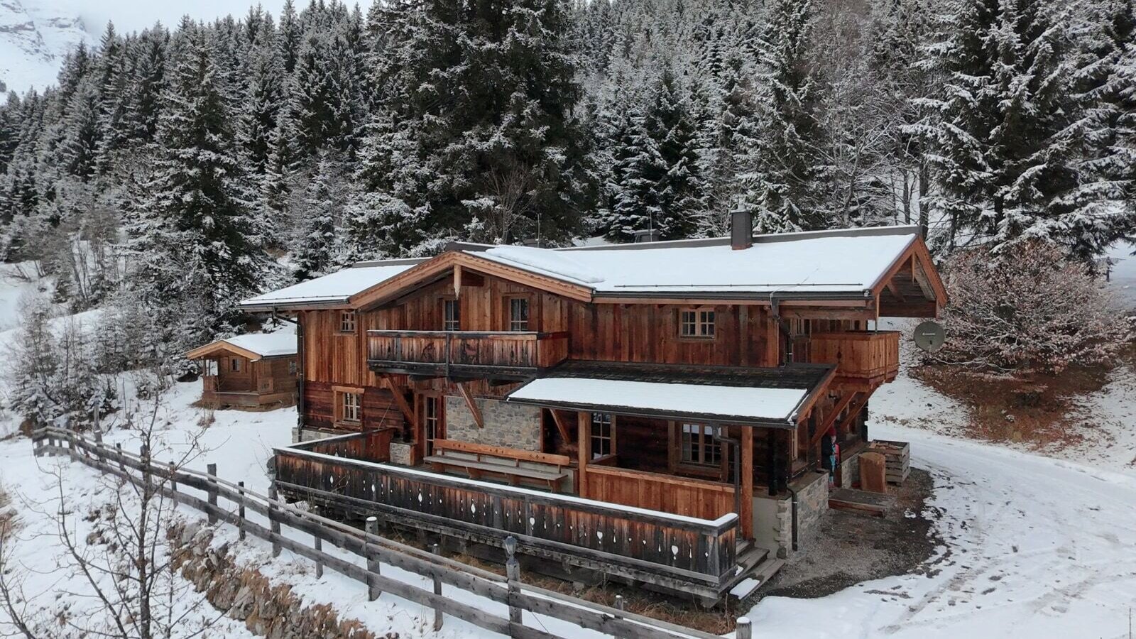 Die verschneite Alm von außen, ein Holzgebäude mit Balkonen, umgeben von Tannen und einem Holzzaun.