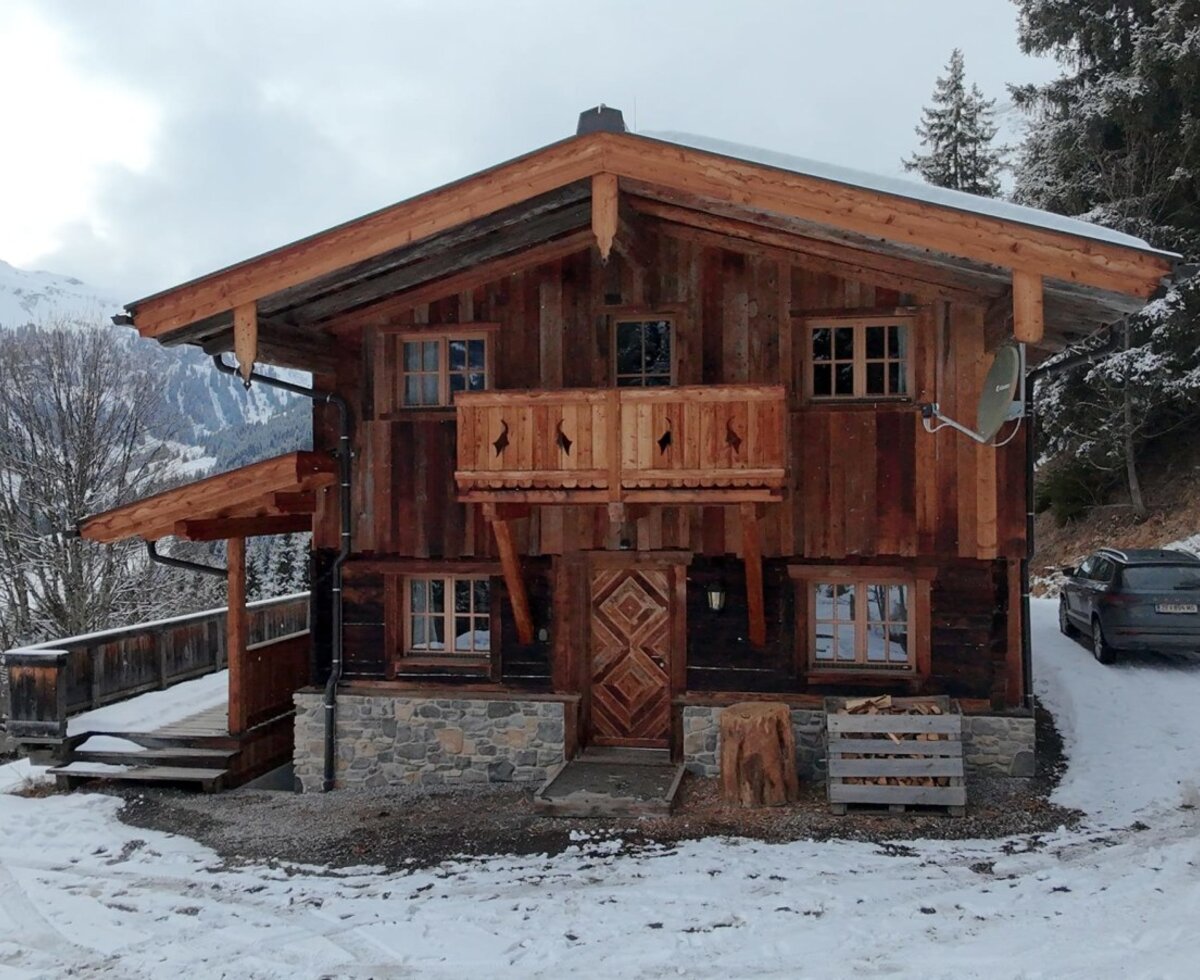 Die Alm, ein Holzhaus mit Steinfundament, Balkon und Parkplatz, umgeben von verschneiten Bergen und Bäumen.