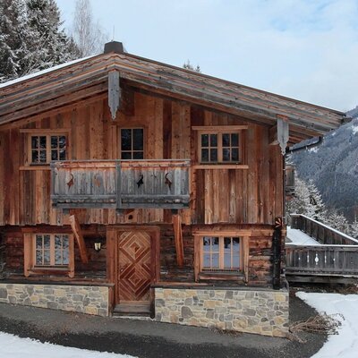 Die Alm im Winter, mit Holzfassade, Steinsockel, Balkon und einer verschneiten Berglandschaft im Hintergrund.