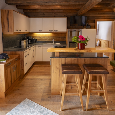 Die Küche der Alm mit einer voll ausgestatteten Küchenzeile, einer Frühstücksbar mit zwei Hockern und einem angrenzenden Wohnbereich mit Sofa.