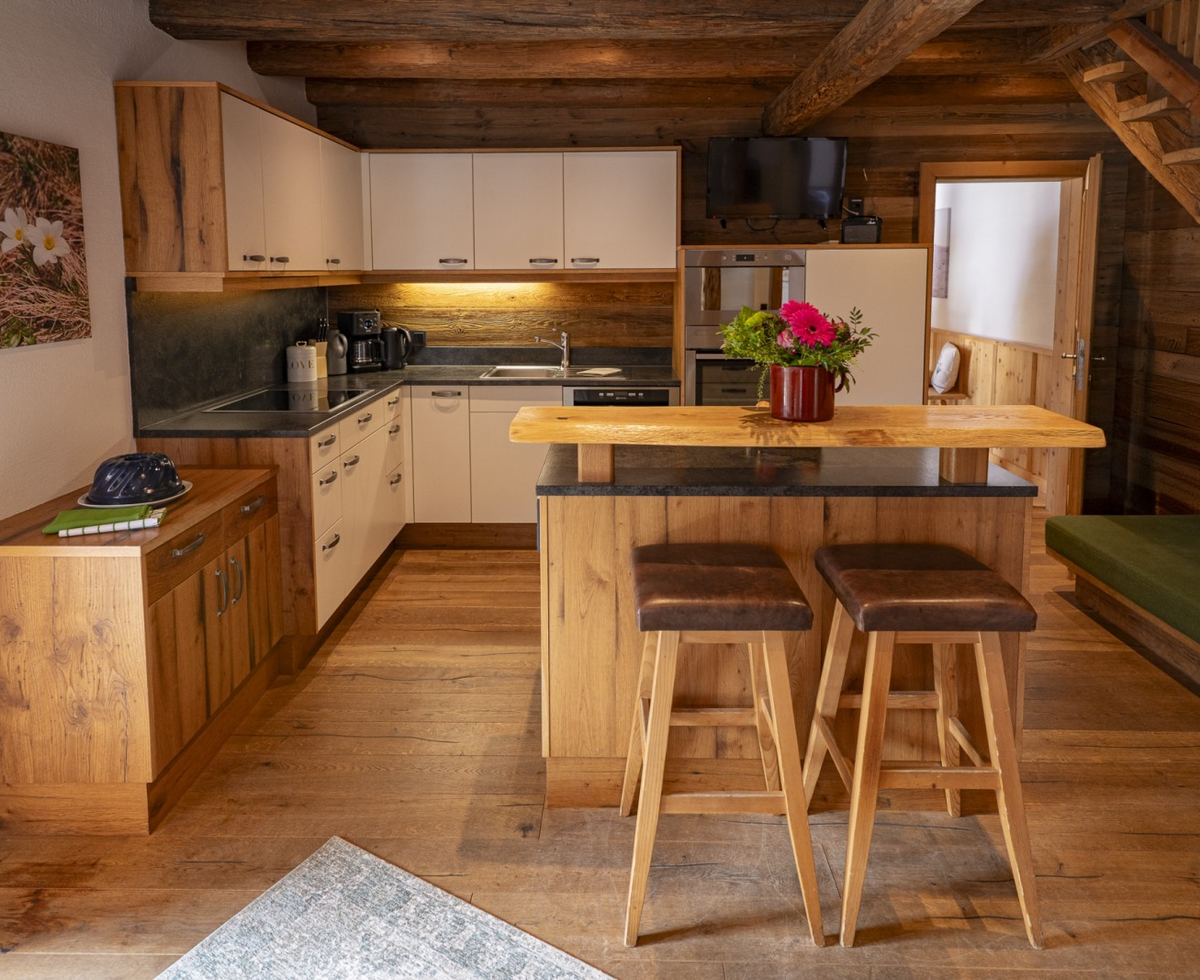 Die Küche der Alm mit einer voll ausgestatteten Küchenzeile, einer Frühstücksbar mit zwei Hockern und einem angrenzenden Wohnbereich mit Sofa.