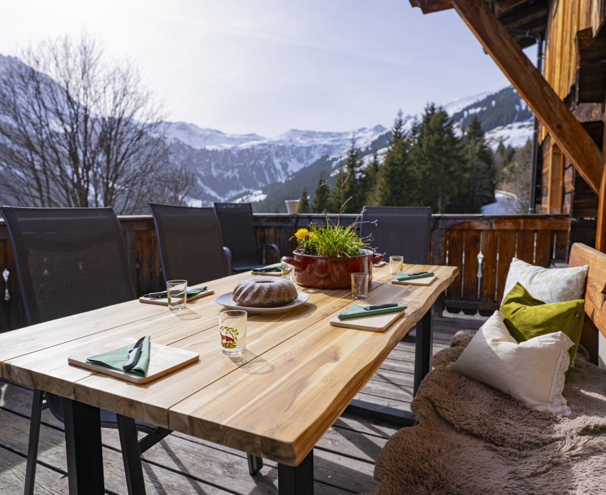 Der Balkon der Alm mit einem gedeckten Holztisch, Sitzgelegenheiten und Blick auf die verschneiten Berge.