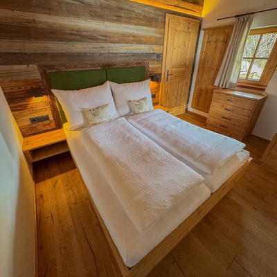Schlafzimmer in der Alm mit Doppelbett, Holzwänden, Nachttischen, Kommode und einem Fenster.