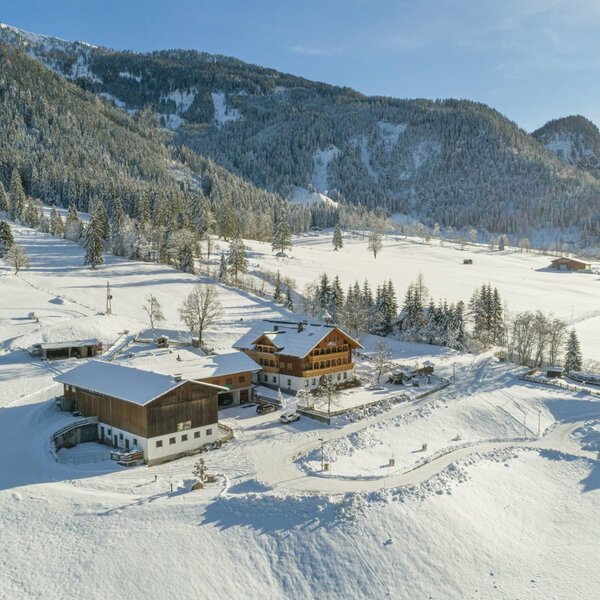 Der Bauernhof in einer verschneiten Winterlandschaft, umgeben von Bergen und Wäldern.