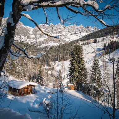 Die winterliche Umgebung des Hofes mit einer schneebedeckten Holzhütte, Tannenbäumen und Bergen im Hintergrund.