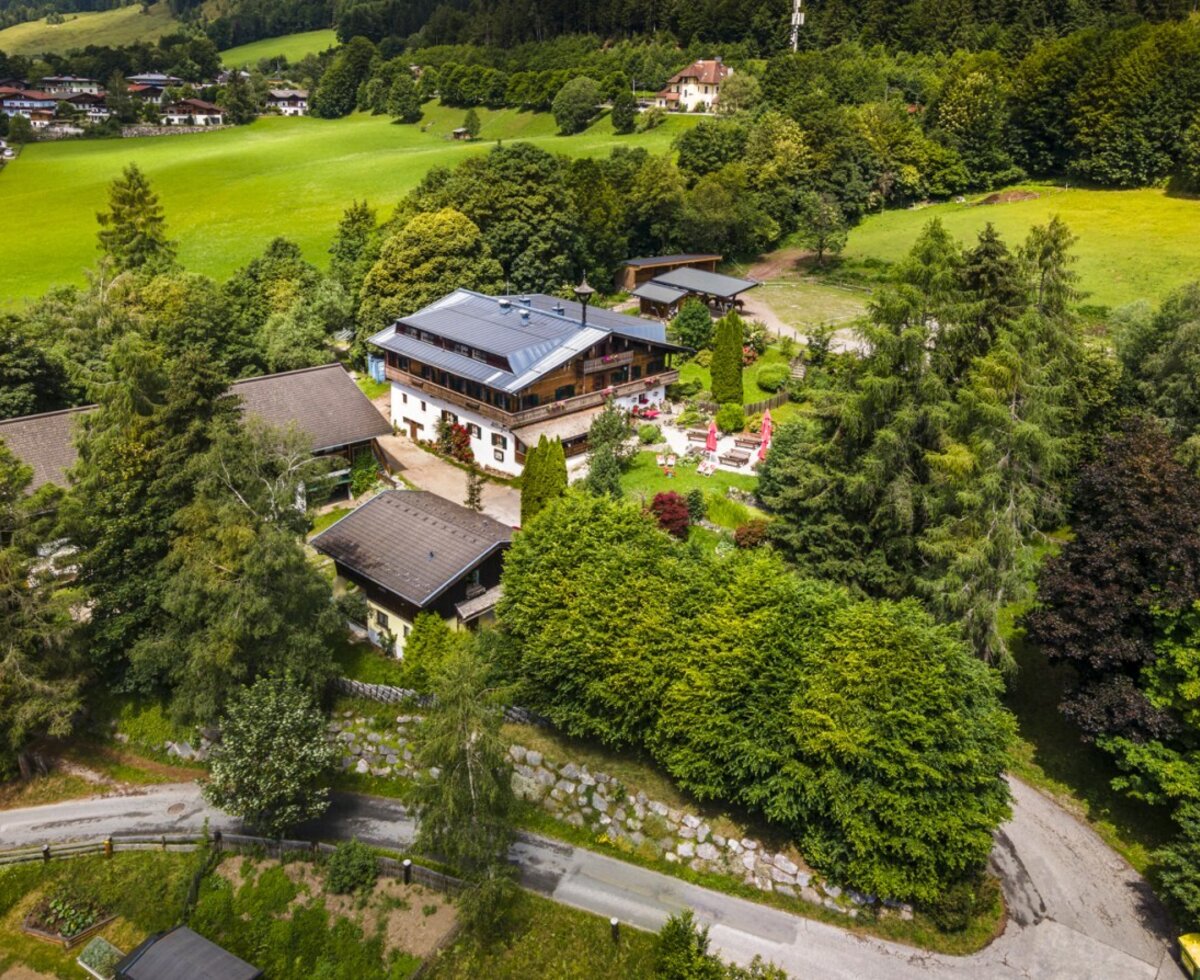 Vogelperspektive auf den idyllischen Bauernhof, umgeben von dichten Wäldern und saftig grünen Wiesen.