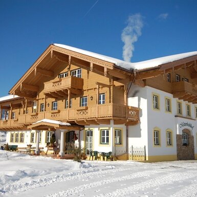 Der winterliche Bauernhof mit Holzbalkonen, schneebedecktem Dach und rauchendem Schornstein, vor der Kulisse verschneiter Berge.