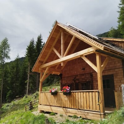 Die Mosermandlhütte mit der neuen überdachten Holzterrasse und Blumenkästen, umgeben von grüner Berglandschaft.