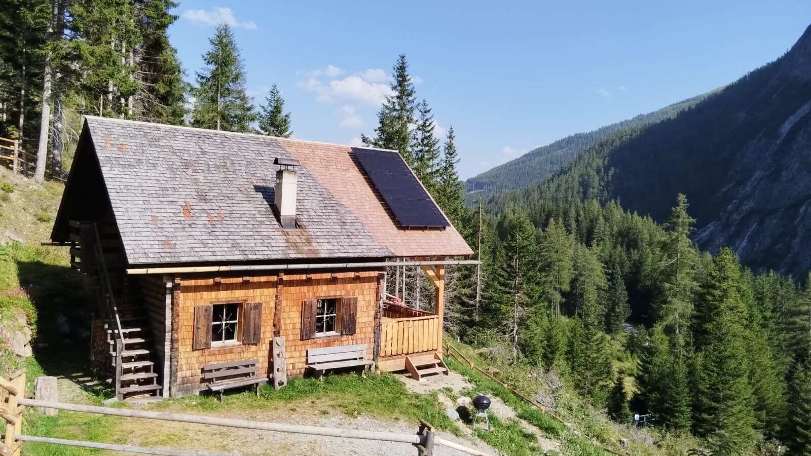 Die Hütte, umgeben von Wald und Bergen, mit Solarmodulen auf dem Dach und Holzbänken vor dem Eingang.
