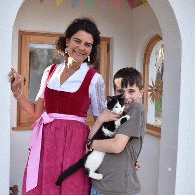 Die Gastgeberfamilie des Bauernhofs heißt Gäste willkommen, dargestellt durch eine Frau in Tracht und ein Kind mit Katze.