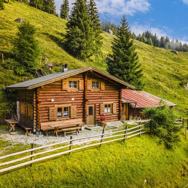 Der Hof in Holzbauweise liegt an einem grünen Hang mit Bäumen und verfügt über einen Außensitzbereich mit Holzbänken.