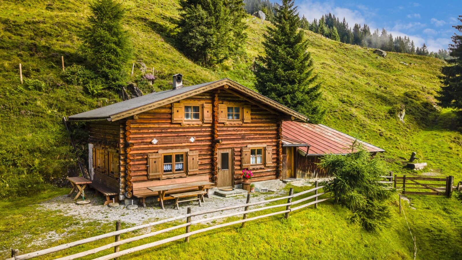 Der Hof in Holzbauweise liegt an einem grünen Hang mit Bäumen und verfügt über einen Außensitzbereich mit Holzbänken.