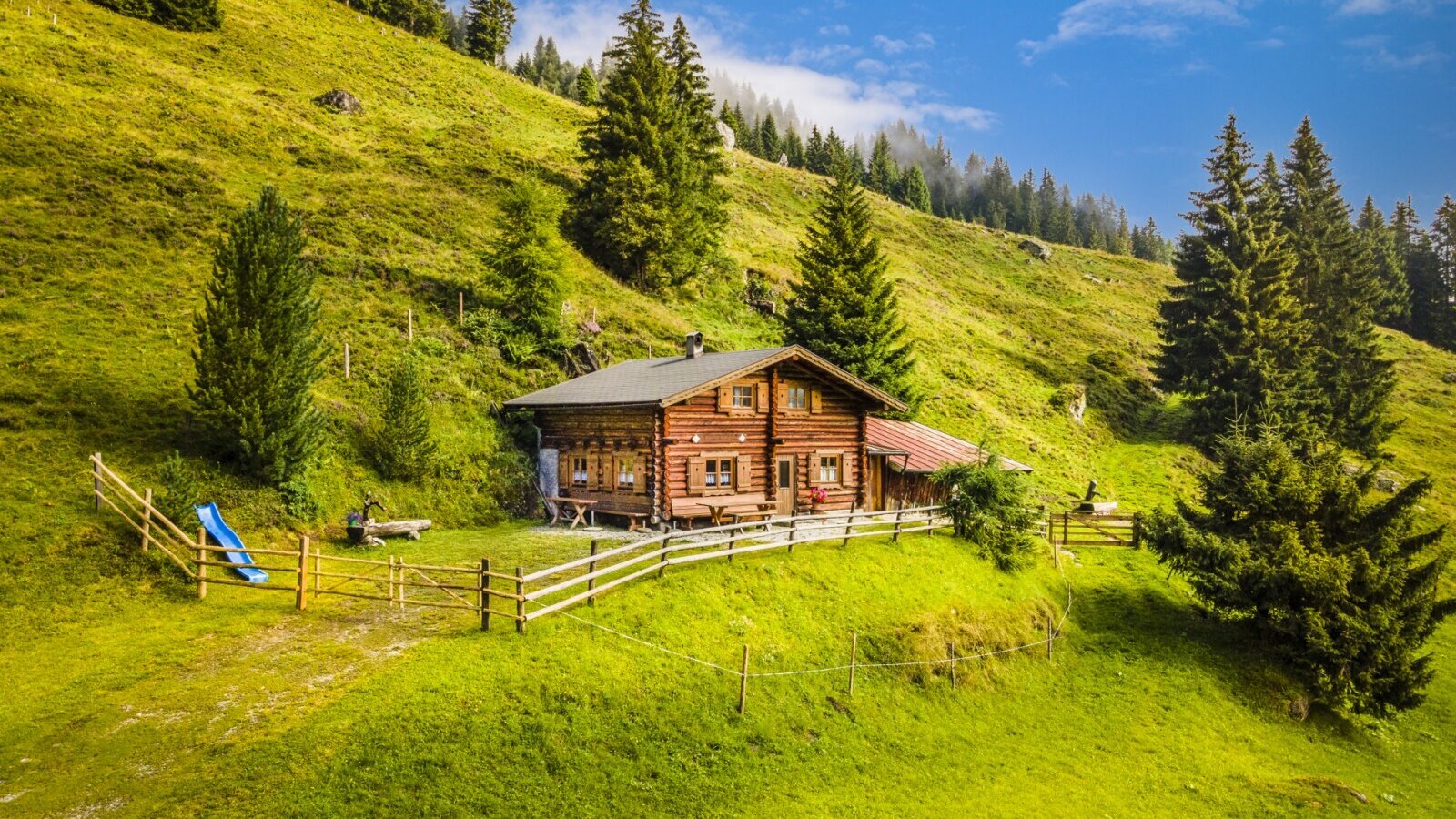 Der Hof liegt auf einer grünen Wiese am Hang, umgeben von Bäumen, mit einem umzäunten Außenbereich und einer Rutsche.