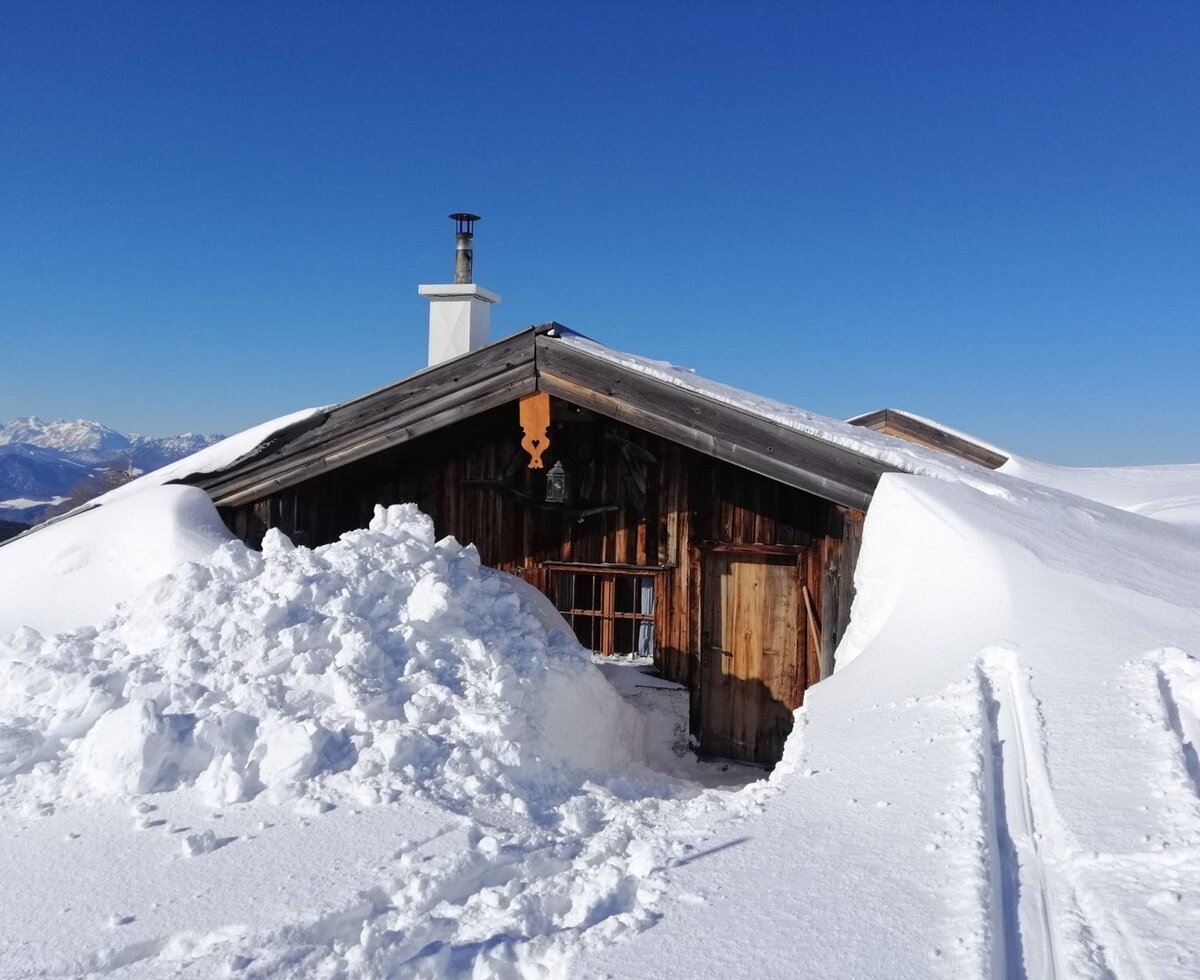 Die verschneite Holzhütte auf dem Hof mit Kamin, tiefem Schnee und Skispuren vor einer Bergkulisse.