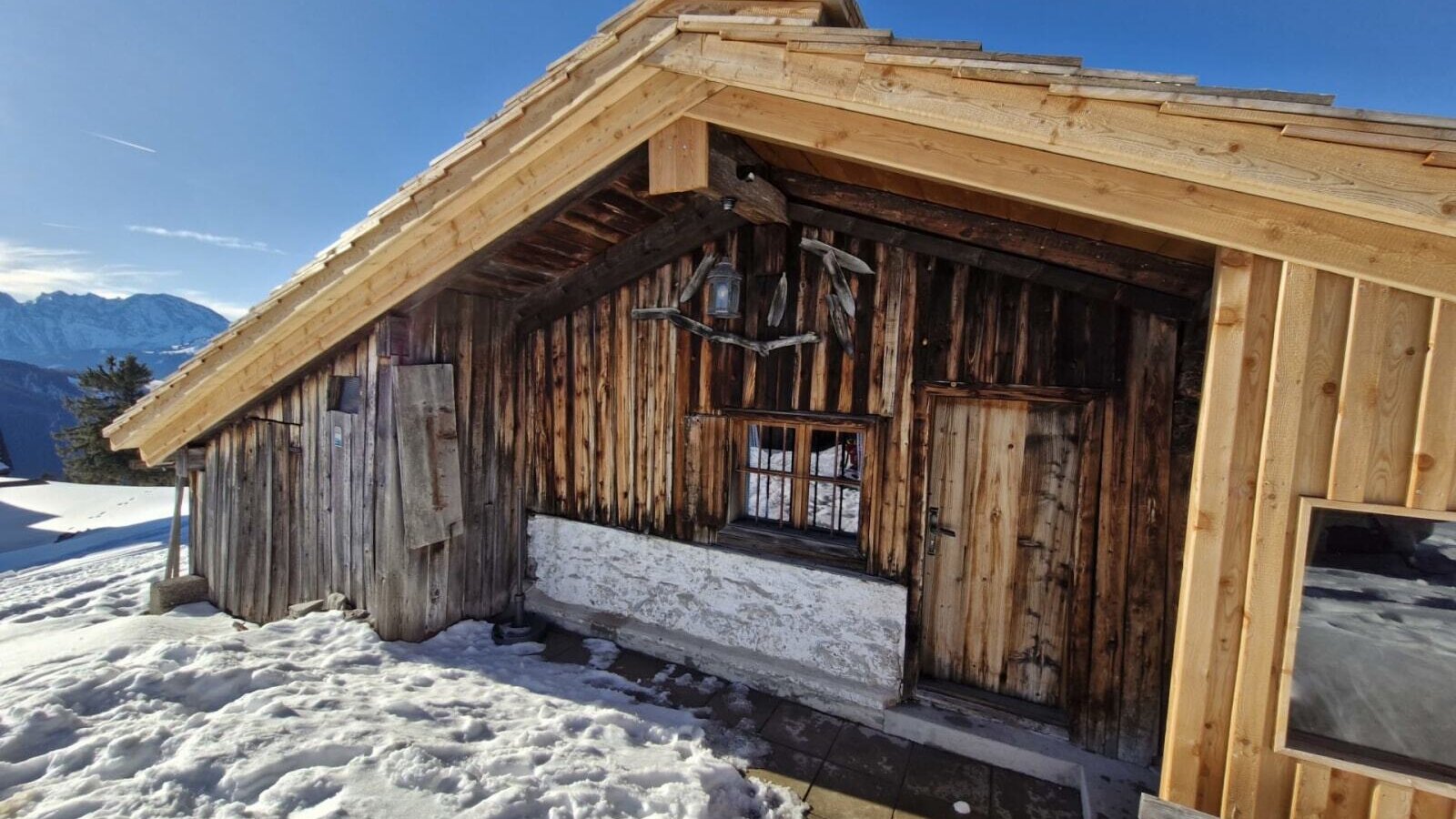 Außenansicht der Hütte  mit Holzfassade, Holztür und verschneiter Umgebung vor Bergkulisse.