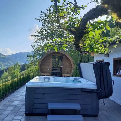 Ein einladender Whirlpool auf der gepflasterten Terrasse der Alm mit einer urigen Fasssauna und weitem Blick auf die grünen Berghänge.