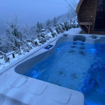 Der einladende Außen-Whirlpool der Alm mit blau beleuchtetem Wasser, umgeben von einer verschneiten Winterlandschaft und einer gemütlichen Fasssauna.