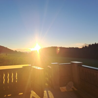 Blick vom Balkon der Ferienwohnung auf den Sonnenaufgang über Feldern, Wald und Bergen.