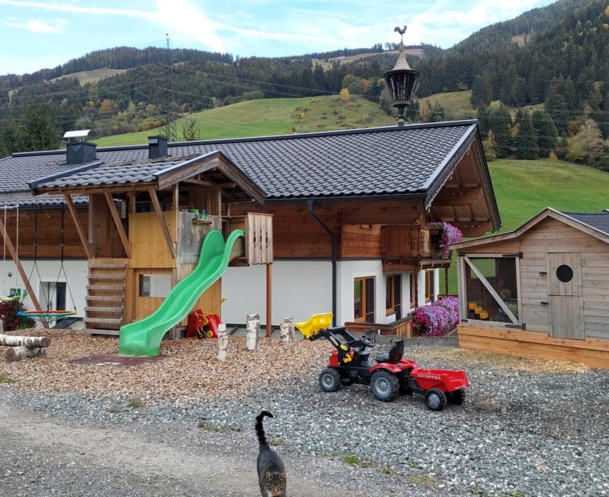 Der einladende Spielbereich des Bauernhofs mit Rutsche, Schaukeln und einem kleinen Spielhaus vor einer malerischen Bergkulisse.