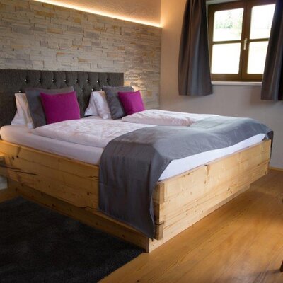 Doppelzimmer im Bauernhof mit Holzbett, gepolstertem Kopfteil, Steinwand, indirekter Beleuchtung und Fenster mit Vorhängen.