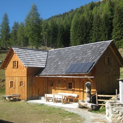 Der Hof ist ein Holzgebäude mit Terrasse, Außensitzgelegenheiten und einem Solarpanel auf dem Dach, gelegen vor einer Waldkulisse.