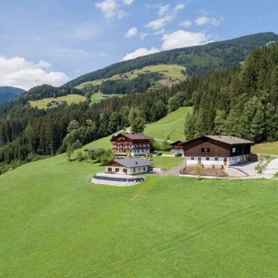 Der Bauernhof mit seinen Gebäuden, umgeben von grünen Wiesen, bewaldeten Bergen und Heuballen.