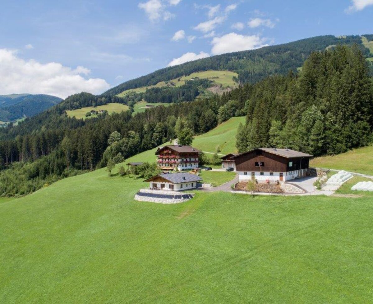 Der Bauernhof mit seinen Gebäuden, umgeben von grünen Wiesen, bewaldeten Bergen und Heuballen.