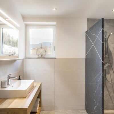 Modernes Badezimmer mit Waschbecken auf Holzunterschrank, beleuchtetem Spiegel, begehbarer Dusche und Fenster mit Bergblick.