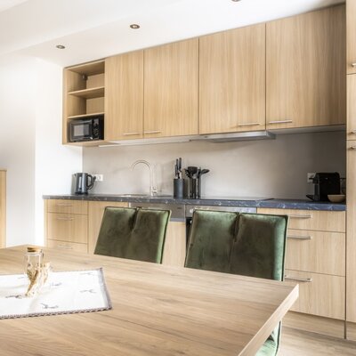 Moderne Küche mit Holzelementen, Esstisch, grünen Stühlen und Küchengeräten wie Mikrowelle und Kaffeemaschine.