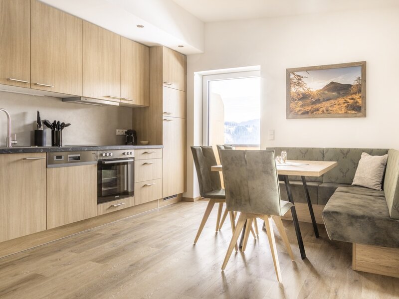 Moderne Küche mit Holzelementen, Backofen, Spülmaschine und Essbereich mit Eckbank und Blick nach draußen.