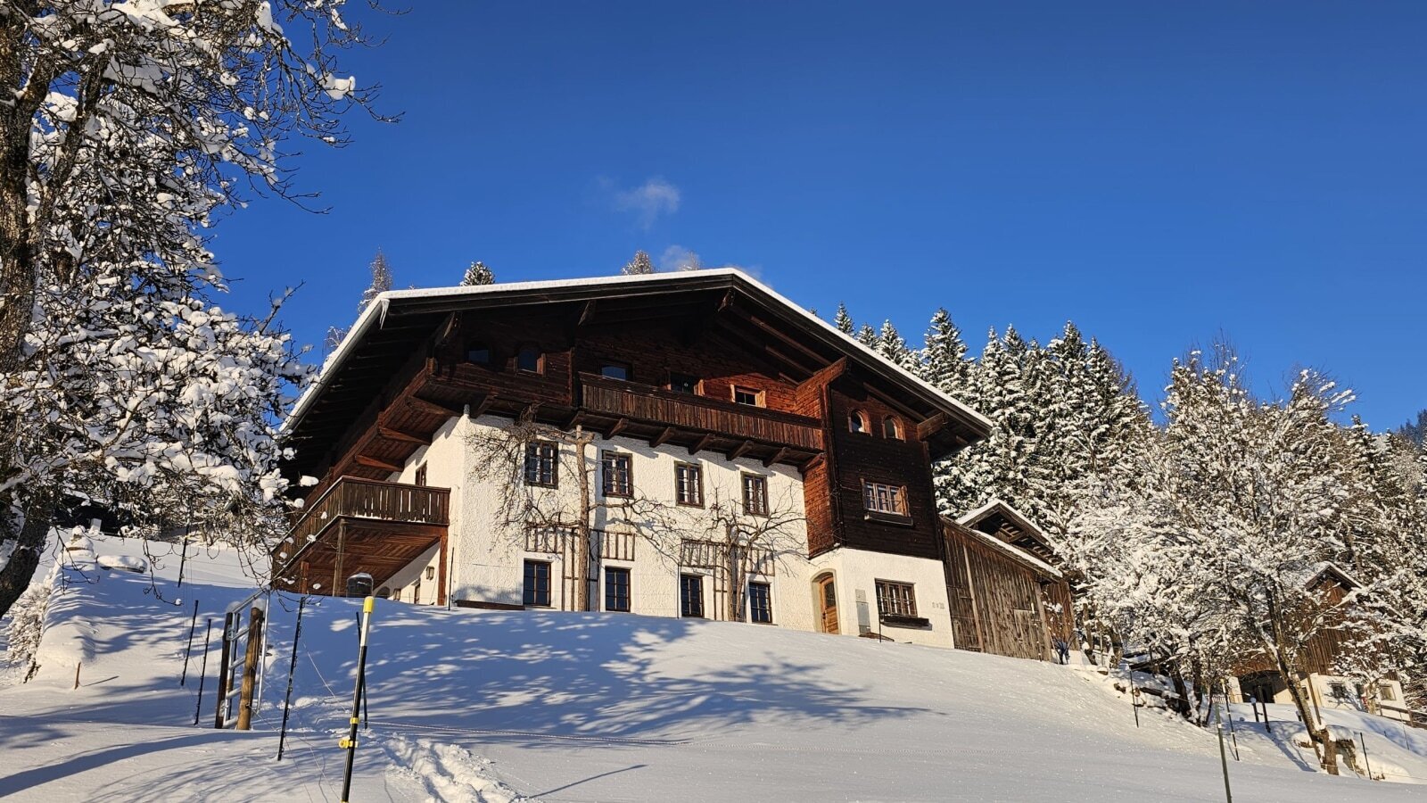 Der Bauernhof in einer winterlichen Landschaft mit Schnee und verschneiten Bäumen unter klarem blauem Himmel.